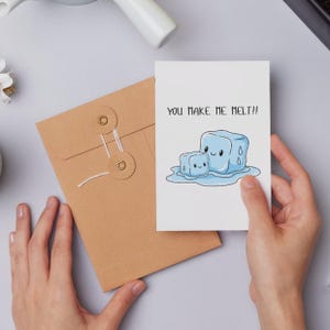 Puede incluir: Una tarjeta blanca con el texto "YOU MAKE ME MELT!!" y una ilustración de dos cubitos de hielo de dibujos animados derritiéndose. La tarjeta está en manos de una persona, junto a un sobre marrón.