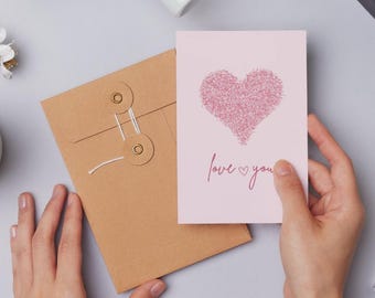 May include: A pink greeting card with a glittery heart design and the words "love you" in cursive. The card is held by a person's hands, next to a brown envelope with string closure.