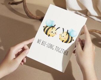 May include: A white greeting card featuring two cartoon bees with black and yellow stripes and blue wings. The text "WE BEE-LONG TOGETHER" is printed below the bees. The card is held by two hands.