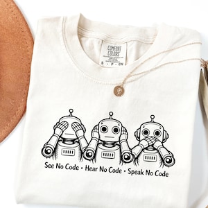 May include: Cream-colored t-shirt featuring three cartoon robots in a "See No Evil, Hear No Evil, Speak No Evil" pose. The robots are black and white, with the text "See No Code • Hear No Code • Speak No Code" below them. A gold necklace is draped on the shirt.