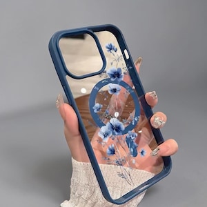 Könnte beinhalten: Eine transparente Handyhülle mit marineblauem Rahmen und floralem Design. Die Hülle verfügt über einen Ringhalter mit blauen Blumen und eine transparente Rückseite. Das Blumenmuster umfasst blaue Blumen und grüne Stiele.