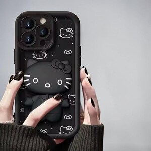 May include: Black phone case featuring a 3D Hello Kitty design. The case has a raised, textured design of the iconic character with a bow, along with smaller Hello Kitty outlines and bows. The case is held by a person with black nail polish.