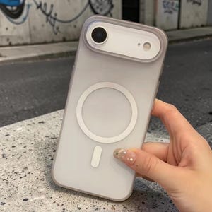 Könnte beinhalten: Eine transparente Handyhülle mit einem weißen kreisförmigen Design und einem kleinen rechteckigen Design. Die Hülle hat einen hellvioletten Rand um die Kamera und die Kanten des Telefons. Die Handyhülle wird von einer Hand gehalten.