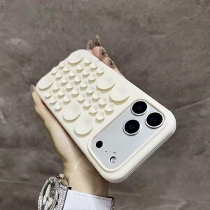 Könnte beinhalten: Eine cremefarbene Smartphone-Hülle mit einem Raster aus zahlreichen kleinen, kreisförmigen Saugnäpfen auf der Rückseite. Die Hülle verfügt über einen rechteckigen Ausschnitt für ein Dreifach-Kamerasystem und einen Blitz sowie eine matte Oberfläche.