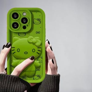 May include: A bright green phone case featuring a 3D Hello Kitty design. The case has a raised outline and includes the camera cutouts. The design includes Hello Kitty's face, bow, and other decorative elements. The case is held by a person.