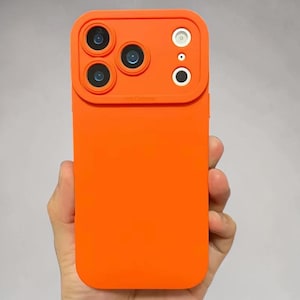 Puede incluir: Funda de teléfono naranja brillante con protector de lente de cámara. La funda tiene un diseño rectangular con una sección elevada para las lentes de la cámara. El protector de la cámara tiene cuatro aberturas circulares para lentes y dos aberturas circulares más pequeñas.