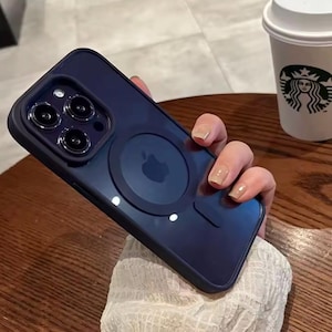 Puede incluir: Funda de teléfono azul marino con borde negro y un accesorio magnético circular. La funda tiene una parte trasera transparente, revelando el diseño del teléfono. El logotipo de Apple es visible.