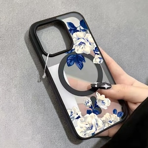 May include: Clear phone case with a black border, featuring a floral design of white roses and blue leaves. The case has a black circular element and a clear back.