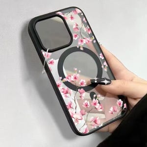 Könnte beinhalten: Transparente Handyhülle mit schwarzem Rahmen und floralem Design mit rosa Kirschblüten. Die Hülle hat einen schwarzen Magnetring und einen transparenten Kameraausschnitt.