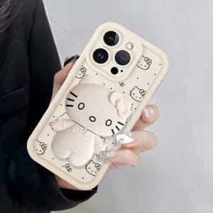 May include: A cream-colored phone case featuring a 3D Hello Kitty design. The case has a raised outline of Hello Kitty, with black details for the eyes, nose, and whiskers. Small Hello Kitty faces are printed across the surface.