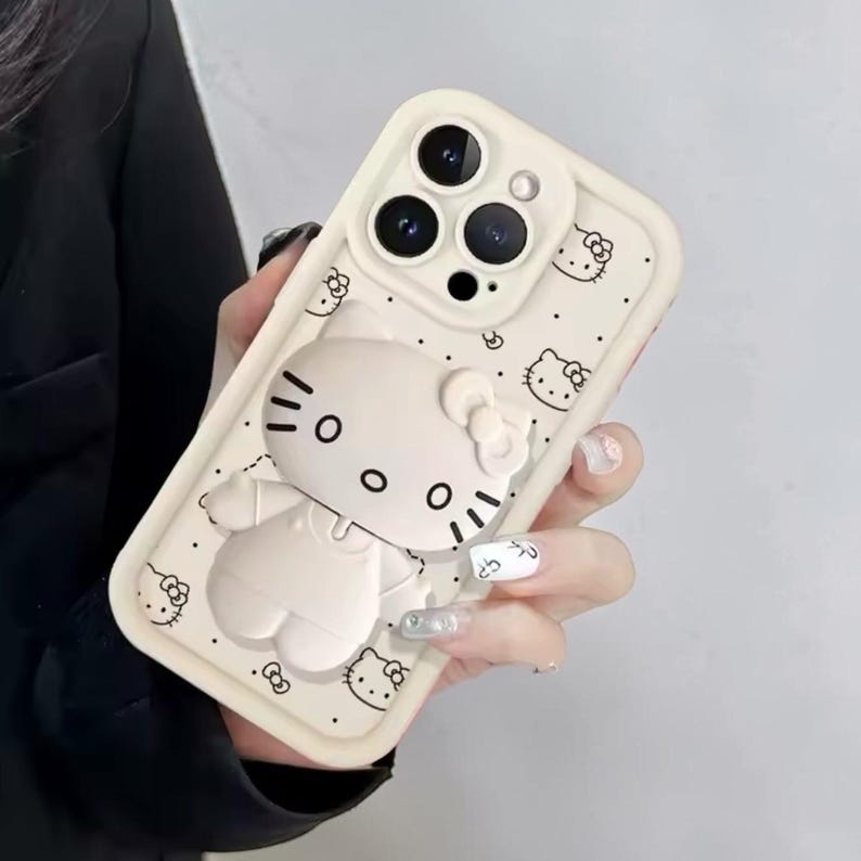 May include: A cream-colored phone case featuring a 3D Hello Kitty design. The case has a raised outline of Hello Kitty, with black details for the eyes, nose, and whiskers. Small Hello Kitty faces are printed across the surface.