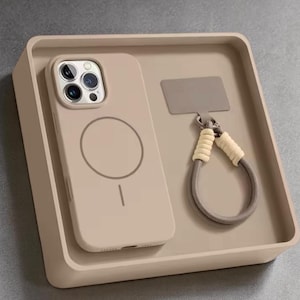 Könnte beinhalten: Eine beige Handyhülle mit kreisförmigem Design und passender Handschlaufe, präsentiert in einer quadratischen Ablage. Die Hülle hat einen Magnetkreis und eine Linie unten. Die Handschlaufe hat einen beigen Griff und ein graues Kabel.