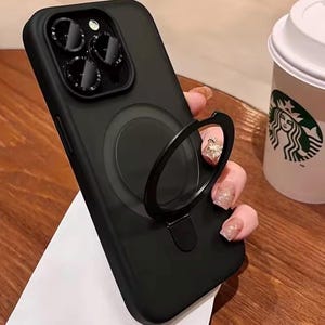 May include: Black phone case with a ring holder. The case has a circular magnetic attachment and a raised camera lens area. A white paper and a Starbucks cup are in the background.