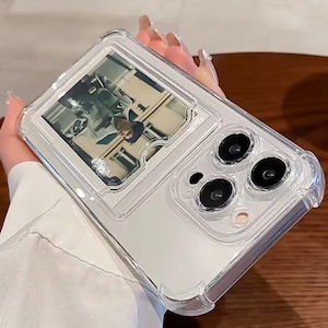 Op de afbeelding: Transparante telefoonhoes met een foto-inzet. De hoes heeft een transparante, beschermende bumper en een rechthoekige sleuf voor een foto. De cameralenzen van de telefoon zijn zichtbaar. De foto toont een persoon.