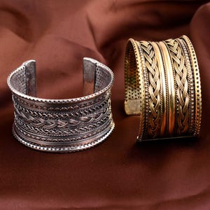 May include: Two wide cuff bracelets, one in a silver-toned metal and one in a gold-toned metal. Both bracelets feature intricate braided patterns, horizontal textured bands, and perforated edges along the top and bottom of the cuffs.