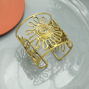 Op de afbeelding: Goudkleurige manchetarmband met een open zonontwerp. De armband heeft een centrale spiraal en stralende lijnen, met bloemmotieven. De sieraden worden gepresenteerd op een lichtgrijze ondergrond.