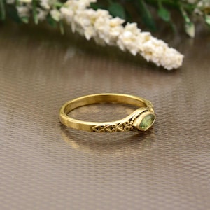 Pode incluir: Um anel de ouro com uma pedra preciosa verde clara. O anel tem um design intrincado na banda e uma moldura para a pedra preciosa. O anel é exibido em uma superfície marrom.