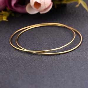 Op de afbeelding: Twee delicate, goudkleurige oorringen worden getoond op een donkergrijze ondergrond. De oorbellen zijn dun en cirkelvormig, met een eenvoudig, elegant ontwerp. De achtergrond toont een vage bloemstuk.