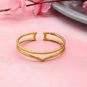 May include: A gold-toned open cuff bracelet featuring a double-band design that converges into a V-shape at the centre. The bracelet has a smooth, polished finish and a gap at the back, allowing for an adjustable fit around the wrist or arm.