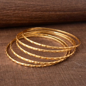 May include: A set of six gold-colored bangles with a textured, geometric pattern. The bangles are stacked on a wooden surface, showcasing their design and color. The jewelry is suitable for everyday wear or special occasions.