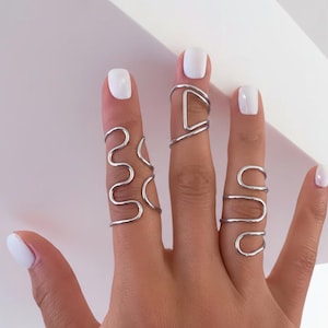 May include: A hand displays five silver-colored, wire-wrapped rings. The rings feature unique designs, including a wavy line, a geometric D-shape, and a series of connected curves. The nails are painted white.