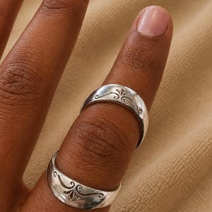 May include: Two polished silver-toned rings worn on a single finger. Each band features a smooth, slightly domed surface with a centered, engraved decorative flourish pattern consisting of symmetrical curved lines and a small central leaf-like motif.