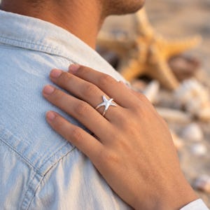 Puede incluir: Un anillo de plata con forma de estrella de mar de cinco puntas usado en un dedo. El anillo presenta un acabado metálico liso y pulido con una banda que se divide en dos brazos finos y curvos que se unen para formar el diseño de estrella.