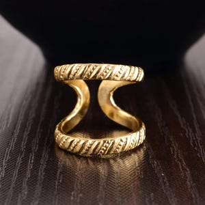 May include: A gold-colored double-band ring with a textured, ridged design. The ring features two bands connected by curved supports, creating a unique and stylish look. The ring is made of a shiny, metallic material.