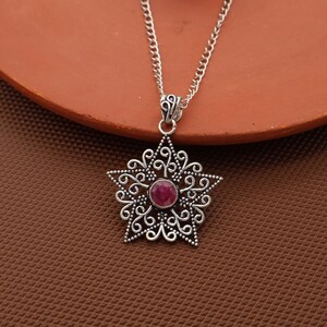 Può includere: Collana argentata con un ciondolo a forma di stella con intricati dettagli in filigrana. Il ciondolo ha una gemma rossa, rotonda e centrale. La catena è a maglia fine e il ciondolo è sospeso da un'ansa decorativa.