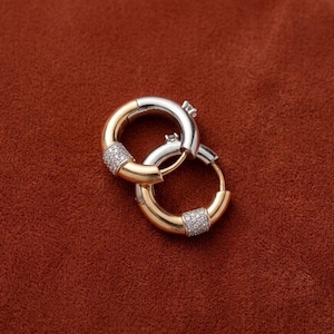 Puede incluir: Dos pendientes de aro unidos. Un pendiente es dorado con una sección de pequeñas piedras transparentes. El otro pendiente es plateado. Ambos pendientes están sobre un fondo marrón rojizo.