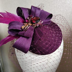 Könnte beinhalten: Ein tiefvioletter Fascinator mit einer großen Schleife, Federn und einem geschmückten Mittelstück. Das Kopfstück hat einen Netzschleier und wird auf einem Schaufensterkopf präsentiert. Die Schleife und die Federn verleihen einen Hauch von Eleganz.