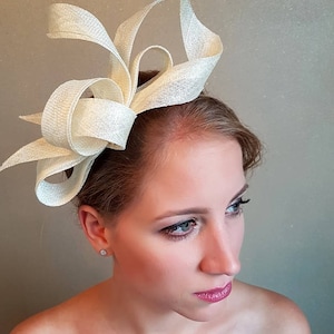 May include: A white, woven fabric hair accessory with a large bow and a curled, feather-like design. The accessory is worn on the side of the head.