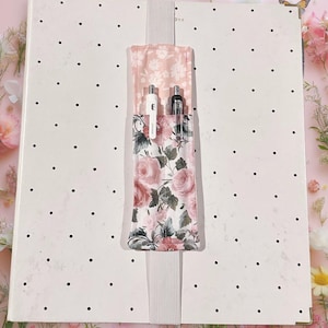 May include: A white notebook with black polka dots and a floral pen holder. The pen holder is made of pink fabric with a rose pattern and holds two pens. A white elastic band secures the notebook.