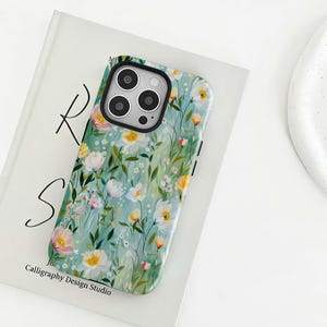 Könnte beinhalten: Eine Handyhülle mit floralem Wiesenmuster mit weißen, gelben und rosa Blumen auf hellblauem Hintergrund. Die Hülle hat ein schwarzes Kameramodul und den Text "Calligraphy Design Studio".