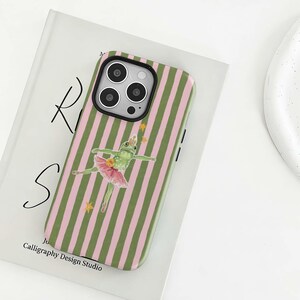 Könnte beinhalten: Handyhülle mit grün-rosa Streifenmuster. Sie zeigt eine Illustration einer Frosch-Ballerina mit Krone und Tutu, mit goldenen Sternen. Die Handyhülle liegt auf einem Buch.