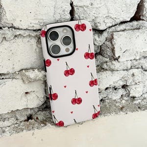 Könnte beinhalten: Weißes Handycase mit einem Muster aus roten Kirschen und kleinen roten Herzen. Das Case hat einen schwarzen Rand um die Kameralinsen und einen schwarzen Rahmen. Der Hintergrund ist eine strukturierte weiße Backsteinmauer.