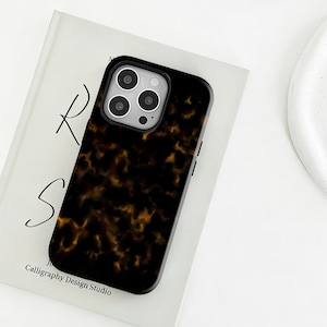 Könnte beinhalten: Eine schwarze Handyhülle mit Schildpattmuster. Die Hülle hat einen schwarzen Rand und einen Kameraausschnitt. Die Handyhülle liegt auf einem weißen Buch mit dem Text "R S" und "Calligraphy Design Studio".