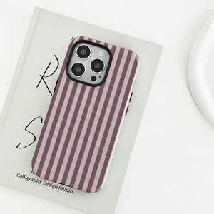May include: A smartphone case featuring a vertical striped pattern in alternating shades of dusty rose and dark mauve. The case has a black camera cutout frame with three circular lens openings and a small flash aperture on the upper left side.