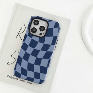 May include: A blue and light blue chequered phone case with a camera cut-out. The case is on top of a book with the text "R S" and "Calligraphy Design Studio". The case has a glossy finish.