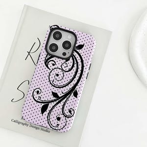 Könnte beinhalten: Eine lavendelfarbene Handyhülle mit einem schwarzen Blumenmuster und kleinen schwarzen Punkten. Die Hülle hat einen schwarzen Kameraschutz und den Text "Calligraphy Design Studio" unten.