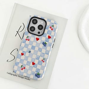 May include: A blue and white chequered phone case featuring red hearts, cherries, and blueberries. The case rests on a book with the text "Ri S" and "Calligraphy Design Studio".