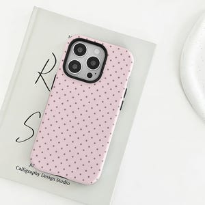 Könnte beinhalten: Eine hellrosa Smartphone-Hülle mit einem sich wiederholenden Muster aus kleinen dunkelgrauen Punkten. Die Hülle hat einen schwarzen Kameraausschnitt mit drei kreisförmigen Objektivöffnungen und einem Blitzsensor.