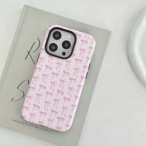 Pode incluir: Uma capa de telefone rosa com um padrão repetido de laços rosa. A capa tem uma borda preta e cobre a parte traseira e as laterais do telefone. A capa do telefone está em cima de um livro com o texto "Calligraphy Design Studio".