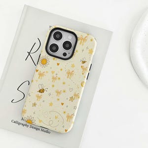 Könnte beinhalten: Cremefarbene Handyhülle mit einem sich wiederholenden Muster aus gelben Schleifen, Bienen, Sternen, Herzen und Blumen. Die Hülle hat einen schwarzen Kamerarahmen und den Text "Calligraphy Design Studio" unten.