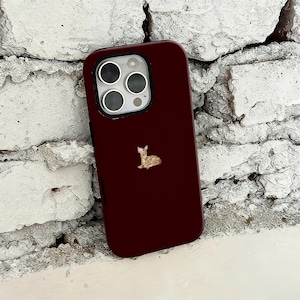 Op de afbeelding: Bordeaux telefoonhoesje met een kleine, gedetailleerde illustratie van een hert. De hoes heeft een glanzende afwerking en een camera-uitsparing met drie lenzen en een flitser. De hertenillustratie is in bruin- en beigetinten.