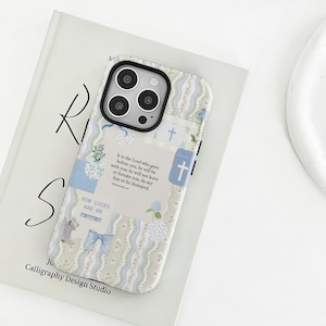 Op de afbeelding: Een telefoonhoesje met een patchwork-ontwerp met bloemmotieven, kruisen en tekst. De hoes heeft een lichtbeige achtergrond met blauwe en witte accenten. De tekst op de hoes luidt "It is the Lord who goes before you..." en "How lucky are we".