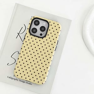 Puede incluir: Una funda para smartphone de color amarillo claro con un patrón repetitivo de pequeños puntos de color marrón oscuro. La funda tiene un recorte rectangular para un sistema de triple cámara y está sobre un libro con el texto Calligraphy Design Studio.