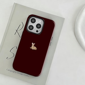 Op de afbeelding: Bordeaux telefoonhoesje met een illustratie van een hert. De hoes heeft een zwarte cameramodule en ligt op een boek met de tekst "Calligraphy Design Studio".