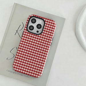 Könnte beinhalten: Eine rot-weiß karierte Handyhülle. Die Hülle liegt auf einem Buch mit dem Text "R S" und "Calligraphy Design Studio". Die Handyhülle hat einen schwarzen Kamerarand.