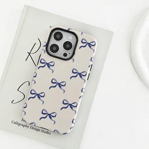 Könnte beinhalten: Eine Handyhülle mit cremefarbenem Hintergrund und einem Muster aus blauen Aquarellschleifen. Die Hülle hat einen schwarzen Rand um die Kameralinsen. Der Text "Calligraphy Design Studio" ist auf dem Buch unter dem Telefon zu sehen.